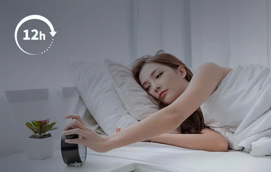 침대에 누워 있는 여성이 알람 시계를 향해 손을 뻗고 있습니다. 흰색 이불에 덮여 있으며, 주변에는 작은 화분이 놓여 있습니다. 이미지의 한쪽에는 '12h'라는 텍스트와 함께 화살표가 그려져 있습니다. 배경은 깔끔하고 밝은 분위기입니다.