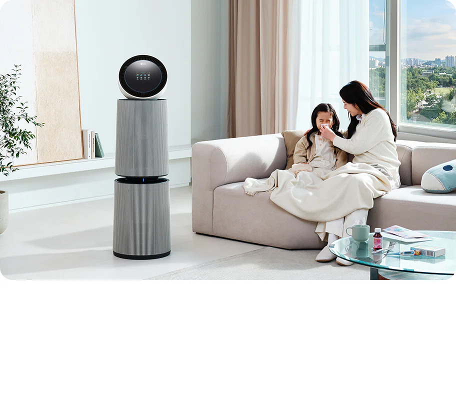 거실 공간에 LG 퓨리케어 AI 오브제컬렉션 360˚ 공기청정기가 놓여 있습니다. 소파 위에는 두 사람이 담요를 덮고 앉아 있으며, 한 여성이 아이의 코를 풀어주고 있는 모습입니다. 테이블 위에는 컵, 약봉투, 약이 놓여 있습니다.